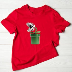 May include: A red t-shirt featuring a graphic of a Piranha Plant from the Super Mario video game series. The plant has a white and red spotted head, green stem and leaves, and is emerging from a green pipe.