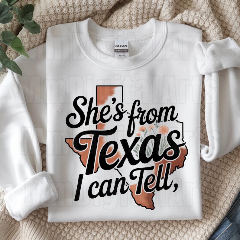 May include: White sweatshirt featuring the phrase "She's from Texas I can Tell," in black script. The design includes a Texas outline with a cowhide pattern and floral accents. The sweatshirt has long sleeves.
