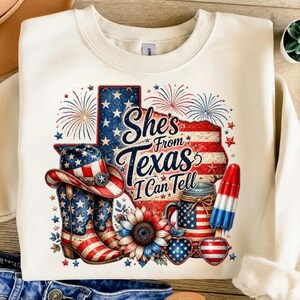 Pode incluir: Uma camisola cor de creme com um gr&aacute;fico de uma bandeira em forma de Texas, fogo de artif&iacute;cio, chap&eacute;u de cowboy, botas, girassol, frasco, gelado e &oacute;culos em forma de cora&ccedil;&atilde;o. O texto diz: She's From Texas I Can Tell.
