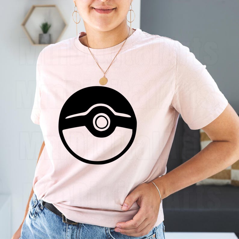 May include: A light pink t-shirt featuring a large black graphic design of a circle with a smaller circle in the center and a horizontal line above and below. The shirt is made of a soft material.