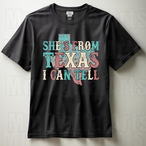May include: Black t-shirt featuring the text "SHE'S FROM TEXAS I CAN TELL" in turquoise, pink, and cream. The design includes a map of Texas. The shirt has a crew neck and short sleeves.