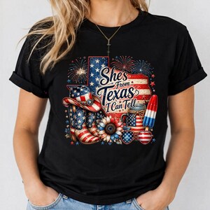 Pode incluir: Uma t-shirt preta com um gr&aacute;fico do estado do Texas com estrelas e riscas. O design inclui uma bota de cowboy, um girassol, um frasco, um gelado e o texto "She's From Texas I Can Tell" com fogo de artif&iacute;cio.