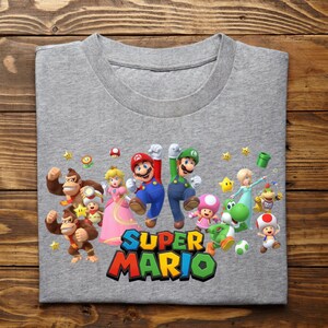 May include: A gray t-shirt featuring a colorful graphic of Super Mario characters, including Mario, Luigi, Princess Peach, and Donkey Kong. The words "Super Mario" are printed in a vibrant, multi-colored font.
