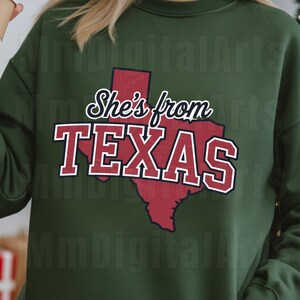 May include: Dark green sweatshirt featuring the text "She's from TEXAS" in white and red, with a red outline of Texas. The sweatshirt is a plain, solid green colour.