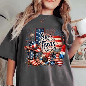 Pode incluir: Uma t-shirt cinzenta escura com um gr&aacute;fico do estado do Texas com padr&otilde;es da bandeira americana, fogo de artif&iacute;cio, um chap&eacute;u de cowboy, botas, um girassol, um frasco e um gelado. O texto diz, She's From Texas I Can Tell.