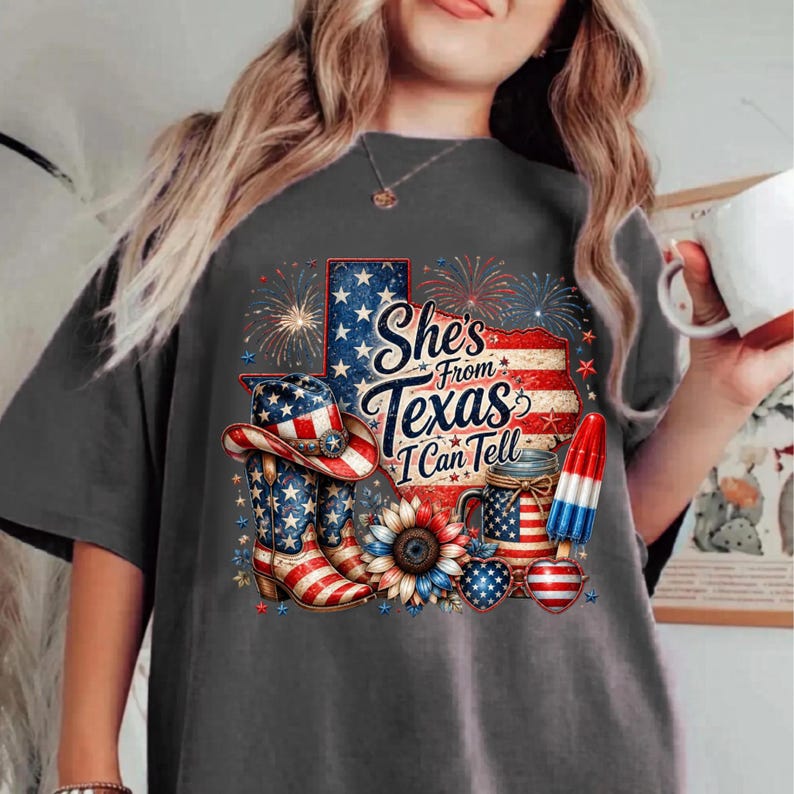 Pode incluir: Uma t-shirt cinzenta escura com um gr&aacute;fico do estado do Texas com padr&otilde;es da bandeira americana, fogo de artif&iacute;cio, um chap&eacute;u de cowboy, botas, um girassol, um frasco e um gelado. O texto diz, She's From Texas I Can Tell.