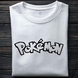 May include: White t-shirt with the word "Pok&eacute;mon" in black, stylized lettering. The shirt has a crew neck and is folded neatly. The background is a dark wood surface.