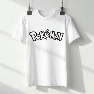 May include: White t-shirt with the word "Pok&eacute;mon" in black, stylized lettering. The shirt is hanging from a metal clip. The shirt has short sleeves and a crew neck.