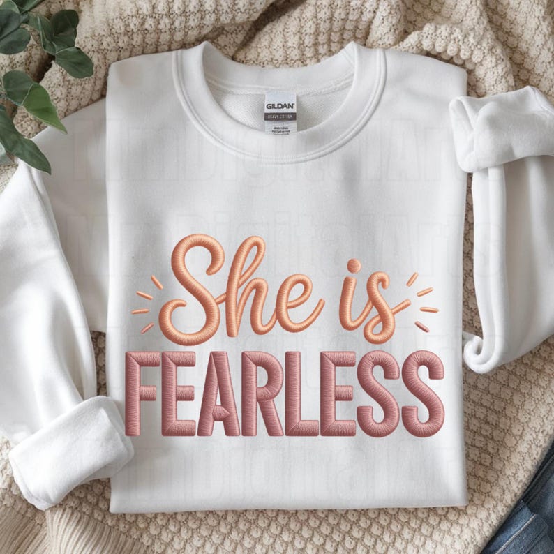 May include: White crewneck sweatshirt with the embroidered text "She is FEARLESS" in a rose gold color. The sweatshirt is folded, with the sleeves rolled up. The Gildan brand tag is visible.