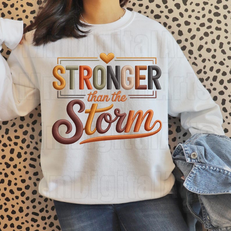 May include: White sweatshirt with the embroidered text "STRONGER than the Storm" in orange, brown, and black. A small gold heart is above the word "STRONGER". The sweatshirt has long sleeves.
