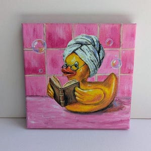 May include: A whimsical painting of a yellow rubber duck wearing a white towel and glasses, reading a book in a pink tiled bathroom. Bubbles float in the air, adding to the playful scene. The duck is the focal point of the artwork.