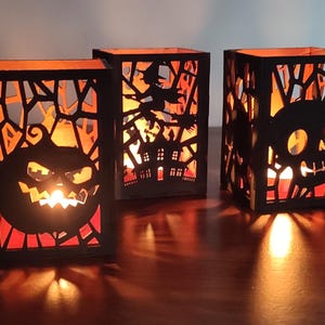 Puede incluir: Tres candelabros negros con temática de Halloween con intrincados diseños recortados. Uno presenta una cara de calabaza, otro una bruja volando sobre una casa embrujada y el tercero una calavera. El cálido resplandor de las velas ilumina los diseños.