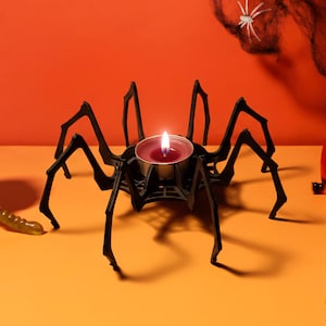Könnte beinhalten: Ein schwarzer, spinnenförmiger Kerzenhalter mit einer brennenden roten Kerze in der Mitte. Die Halloween-Dekoration steht auf einer orangefarbenen Oberfläche, mit einem Totenkopf und einer Kürbislaterne im Hintergrund.