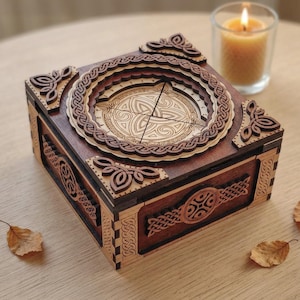 Può includere: Una scatola di legno con intricati disegni a nodi celtici. La scatola è quadrata con una finitura marrone scuro e dettagli in legno più chiaro. Il coperchio presenta un design circolare a strati con un motivo a nodo centrale. I pannelli decorativi angolari e laterali aggiungono all'estetica ornata.