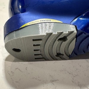 Puede incluir: Primer plano de una suela de zapato gris con un diseño texturizado y una parte superior azul. La suela tiene una serie de ranuras y cortes para la tracción. El texto "anatomic CHASSIS" es visible en el lateral del zapato.