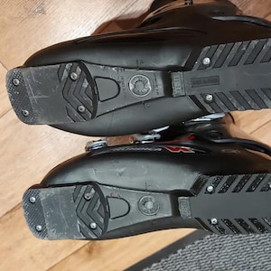 May include: Pair of black ski boots with textured soles and metal hardware. The boots are designed for winter sports and feature a sturdy construction. The soles have a pattern for grip on snow and ice.