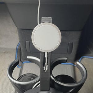 Könnte beinhalten: Ein schwarzer Getränkehalter für das Auto mit einem weißen Ladekabel und einem runden, weißen Ladegerät. Der Getränkehalter ist für die Mittelkonsole eines Autos konzipiert. Das Ladekabel ist an das Innere des Autos angeschlossen.