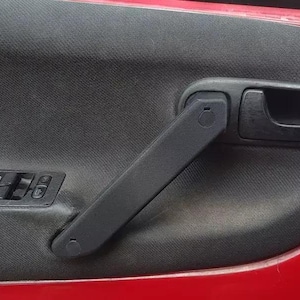 Op de afbeelding: Close-up van een auto-interieur. De afbeelding toont een zwarte deurgreep, een zwart bedieningspaneel voor de ramen en een rood deurframe. Het deurpaneel heeft een getextureerde, donkergrijze stof.