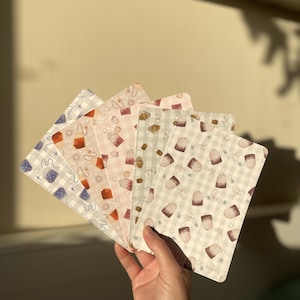 May include: A hand displays a collection of patterned cards. Each card has a unique design, such as tea bags, snowflakes, and geometric shapes, set against a pastel colour scheme. The rectangular cards are likely for journaling or crafting.