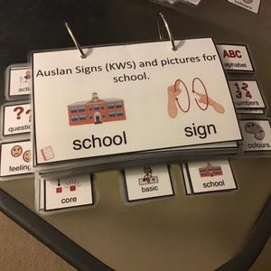 May include: A stack of flashcards with the text "Auslan Signs (KWS) and pictures for school." The cards feature illustrations of a school building and hand signs. Other cards show the alphabet, numbers, and colors.