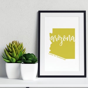 Op de afbeelding: Een ingelijste print met de omtrek van Arizona in een geelgroene kleur, met het woord "arizona" in witte letters. Daaronder staat de tekst "LAND OF SUNSHINE". De zwarte lijst staat op een witte plank met twee potplanten.