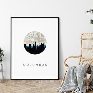 May include: A framed art print featuring a black silhouette of the Columbus, Ohio skyline. The design includes a circular map of the city and the word "COLUMBUS" in a minimalist font. The print is displayed in a room with a chair and a plant.
