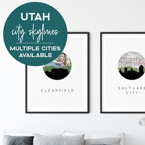 May include: Two framed prints of city skylines with a map background. The prints are titled "Salt Lake City" and "Clearfield". The prints are hanging on a white wall above a bed with grey bedding.