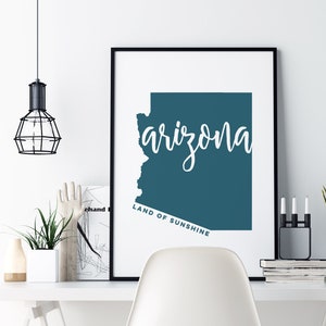 Op de afbeelding: Een ingelijste print met de staat Arizona in teal, met het woord "arizona" in cursief schrift. De tekst "LAND OF SUNSHINE" staat eronder. De print staat op een wit bureau met een witte stoel.