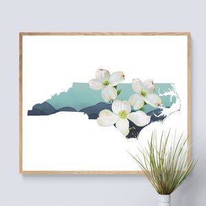 May include: A framed art print featuring the outline of North Carolina in white, with a teal and blue watercolor mountain range design. White dogwood flowers are placed over the state outline. The artwork is in a natural wood frame.
