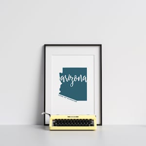 Op de afbeelding: Ingelijste print met de staat Arizona in teal, met het woord "arizona" in witte letters. Daaronder staat "LAND OF SUNSHINE". Een gele typemachine staat voor de ingelijste print.