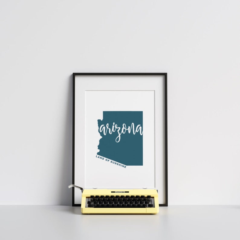 Op de afbeelding: Ingelijste print met de staat Arizona in teal, met het woord "arizona" in witte letters. Daaronder staat "LAND OF SUNSHINE". Een gele typemachine staat voor de ingelijste print.
