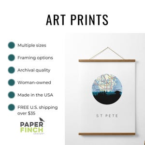 An art print of St. Pete with a circular map and skyline hangs from a wooden magnetic poster hanger on a white wall.