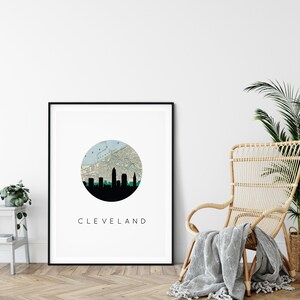 May include: A framed art print featuring a black silhouette of the Cleveland skyline. The design includes a map within a teal circle, with the city name in a clean, modern font below. The print is displayed in a room with a wooden floor and a wicker chair.