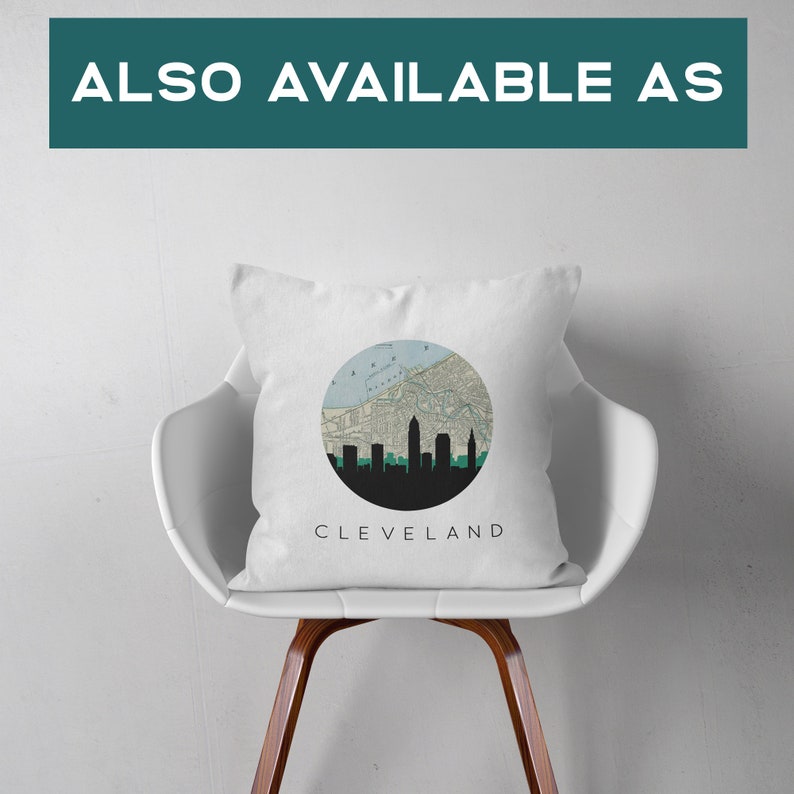 May include: White square pillow with a circular design featuring a map and the silhouette of the Cleveland skyline. The word "CLEVELAND" is printed below the design. The pillow is on a white chair.