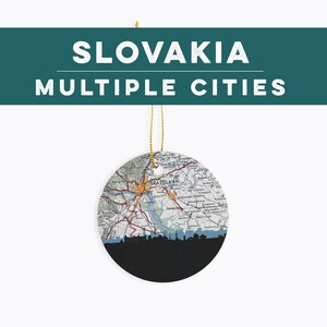 May include: A round ceramic ornament featuring a map of Slovakia with the text "SLOVAKIA MULTIPLE CITIES" above. The bottom half is black with a city skyline silhouette. A gold string is attached for hanging.