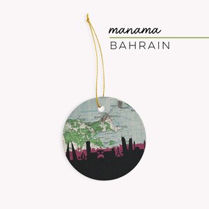 May include: Round ceramic ornament featuring a map of Manama, Bahrain, with a black skyline silhouette at the bottom. The ornament has a gold string for hanging and the words "manama" and "BAHRAIN" are printed on the top.