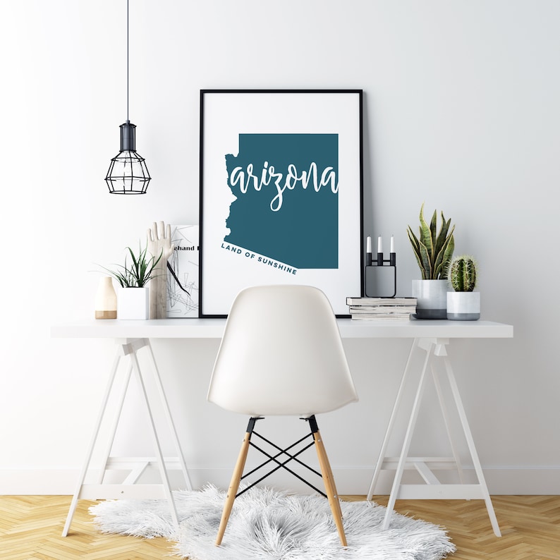 Op de afbeelding: Een ingelijste print met de staat Arizona in teal, met het woord "arizona" in cursief schrift. De tekst "LAND OF SUNSHINE" staat eronder. De print staat op een wit bureau met een witte stoel.