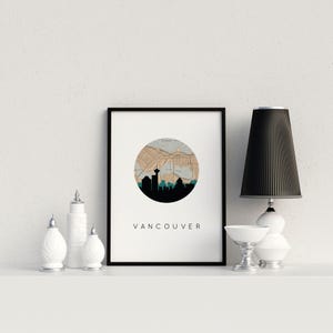 Framed Vancouver skyline art print on a shelf next to small white decorative jars, a small white bowl, and a black-shaded lamp.