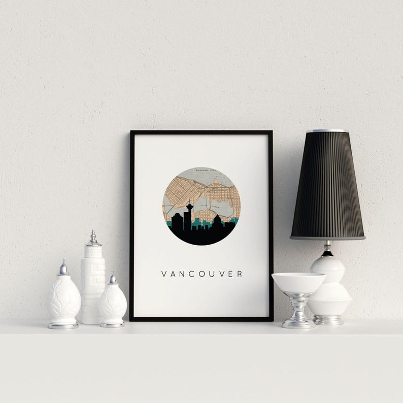 Framed Vancouver skyline art print on a shelf next to small white decorative jars, a small white bowl, and a black-shaded lamp.