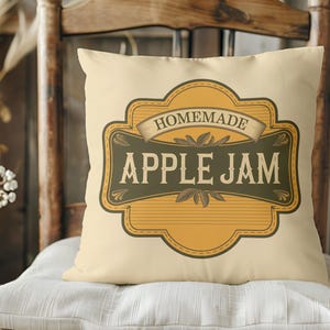 Peut inclure: Coussin crème avec une étiquette décorative indiquant "Homemade Apple Jam". L'étiquette est jaune, marron et verte, avec une bannière en haut. Le coussin est posé sur un coussin blanc, avec une chaise en bois en arrière-plan.