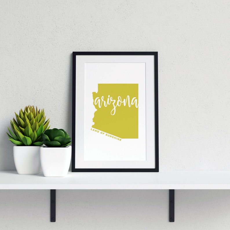 Op de afbeelding: Een ingelijste print met de omtrek van Arizona in een geelgroene kleur, met het woord "arizona" in witte letters. Daaronder staat de tekst "LAND OF SUNSHINE". De zwarte lijst staat op een witte plank met twee potplanten.