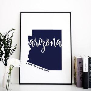 Op de afbeelding: Een ingelijste print met de staat Arizona in een donkerblauwe kleur. Het woord "arizona" is in witte cursieve letters over de omtrek van de staat geschreven. De tekst "LAND OF SUNSHINE" staat eronder in een kleinere, rechte letter.