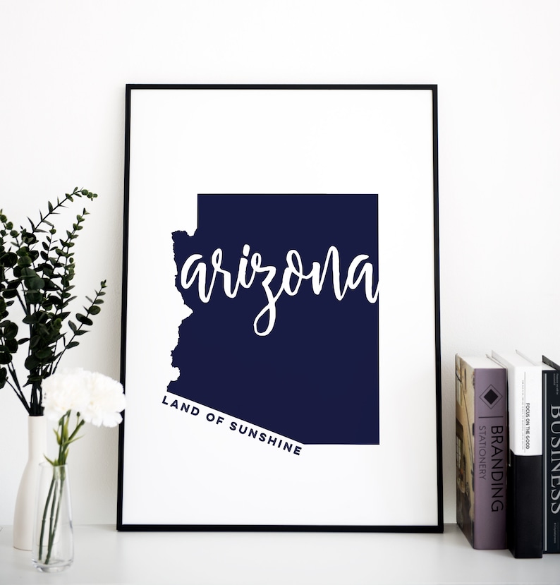 Op de afbeelding: Een ingelijste print met de staat Arizona in een donkerblauwe kleur. Het woord "arizona" is in witte cursieve letters over de omtrek van de staat geschreven. De tekst "LAND OF SUNSHINE" staat eronder in een kleinere, rechte letter.