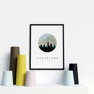 May include: A framed print featuring a black silhouette of the Cleveland skyline set against a map within a circle. The word "CLEVELAND" is printed below. Various colored vases in brown, yellow, pink, blue, and black are arranged in front.