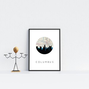 May include: A framed print featuring a black silhouette of the Columbus, Ohio skyline against a white background. The skyline is set within a circular map design. The word "COLUMBUS" is printed in a minimalist font below the image.