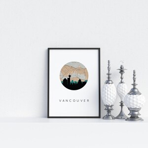 Framed Vancouver art print featuring a circular map and skyline graphic, displayed on a shelf next to three silver decorative objects against a white wall.