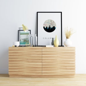May include: A modern interior scene featuring a light wood cabinet with two doors. The cabinet is adorned with decorative items, including a framed print with a city skyline, a yellow lamp, vases, and a book. The word "COLUMBUS" is visible on the print.