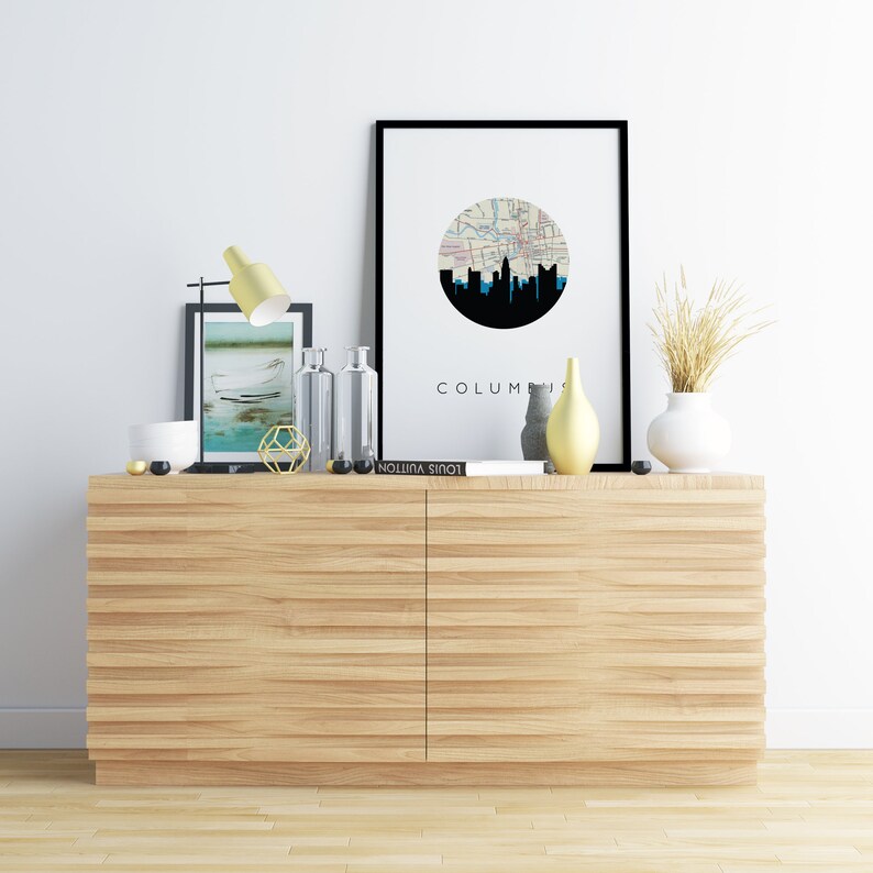 May include: A modern interior scene featuring a light wood cabinet with two doors. The cabinet is adorned with decorative items, including a framed print with a city skyline, a yellow lamp, vases, and a book. The word "COLUMBUS" is visible on the print.