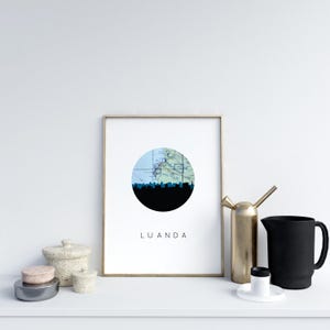 May include: A framed print featuring a silhouette of a city skyline in black, set against a blue and green map within a circular design. The word "LUANDA" is printed in a clean font below the circle. The frame is gold.