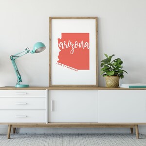 Op de afbeelding: Een ingelijste kunstdruk met de staat Arizona in koraal, met het woord "arizona" in wit script. De tekst "LAND OF SUNSHINE" staat onder de omtrek van de staat. De print staat op een witte en houten kast, met een turquoise bureaulamp en potplanten. De kast is 150 cm breed.